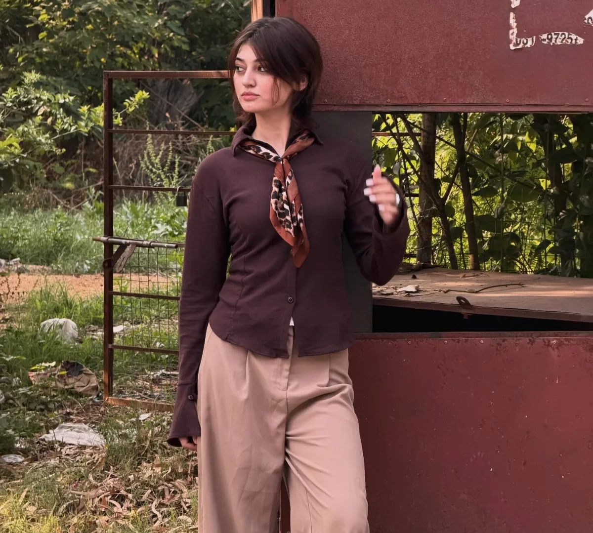 Young woman in a brown shirt, leopard-print neck scarf and beige trousers leaning on a rusty metal booth outdoors