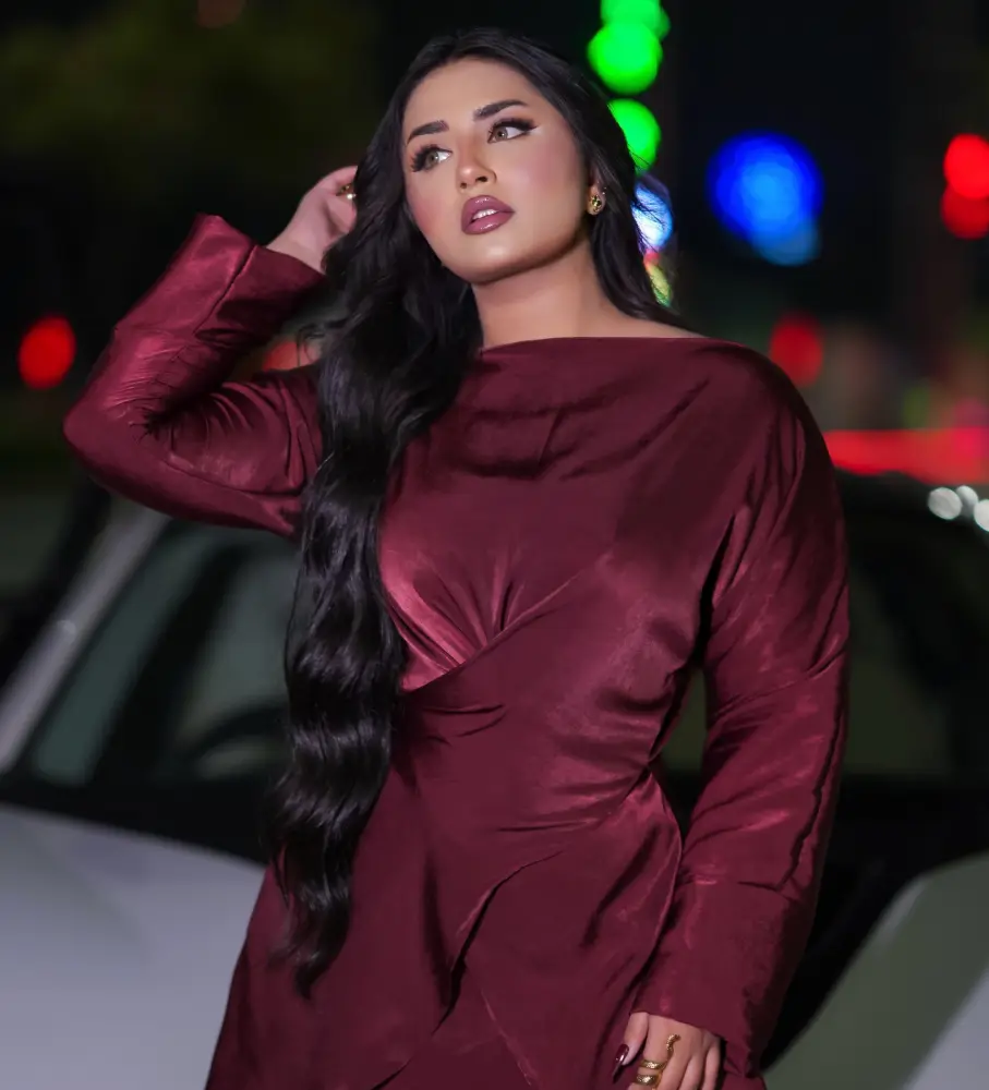 Woman with long dark wavy hair in a burgundy satin dress posing at night near blurred city lights
