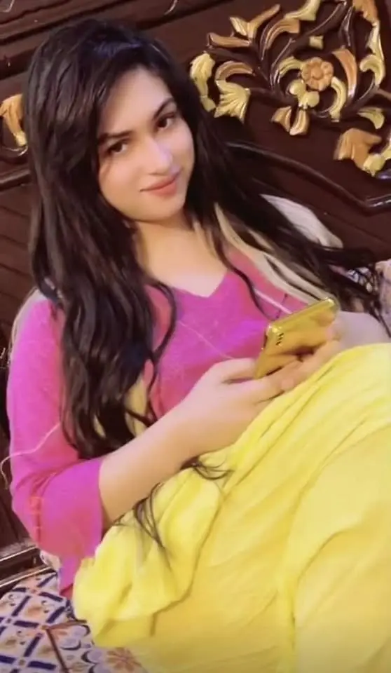 Woman with long dark wavy hair in a pink top and yellow outfit, holding a gold phone before a carved wooden headboard