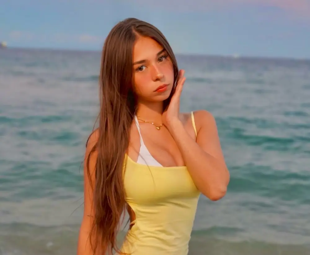 Young woman with long brown hair in a yellow top and gold necklace, touching her cheek while standing in the sea at dusk